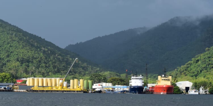 A port facility in Trinidad surrounded by lush green hills, with energy storage tanks, docked ships, and cranes in view—highlighting the infrastructure critical to Southern Caribbean Energy Integration efforts among Trinidad and Tobago, Guyana, and Suriname.