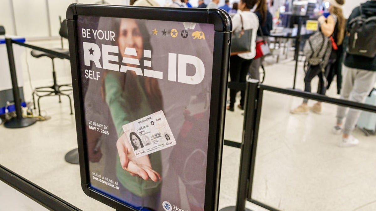 A TSA checkpoint sign at an airport displays a DHS poster promoting REAL ID, showing a woman holding out her ID card with the message