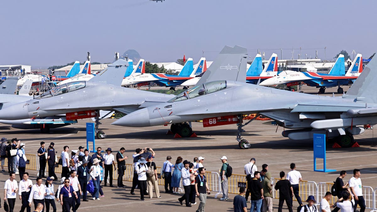 Crowds of visitors walk past several J-15 fighter jets of the Chinese PLA Navy on static display at Airshow China 2024, with other aircraft and a jet flying overhead.