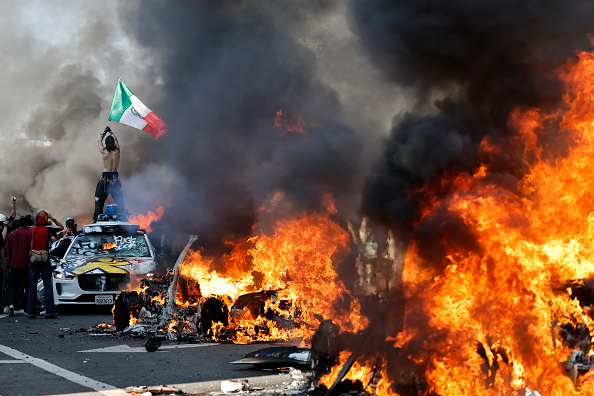 A protestor holds up a Mexican flag as burning cars line the street on June 08, 2025 in Los Angeles, California.
