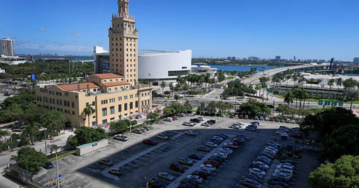 Aerial view of Miami's Freedom Tower alongside a parking lot, with modern buildings and palm trees in the background under a clear blue sky.