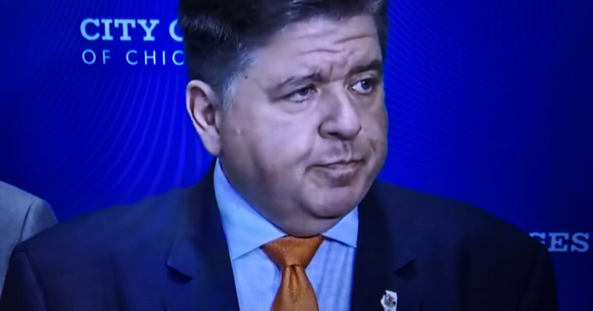 Illinois Governor J.B. Pritzker speaking at a press conference in front of a blue backdrop with the City of Chicago logo.