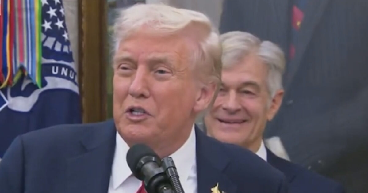 Donald Trump speaking at a podium during an event, with a smiling individual in the background, surrounded by official decorations and flags.