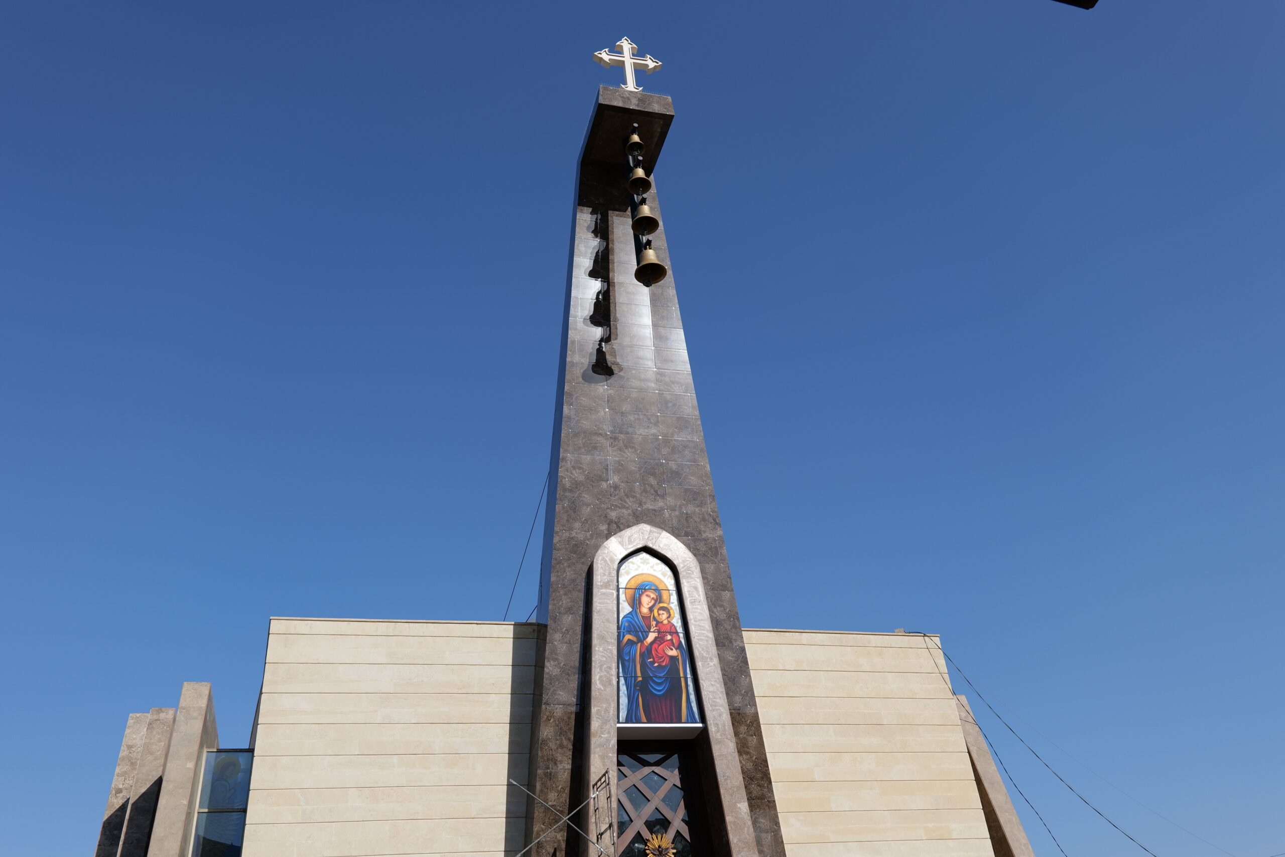 Modern church tower featuring a cross, bells, and an icon of the Virgin Mary against a clear blue sky.