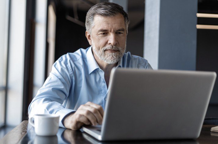 Middle-aged man with a beard working on a laptop in a modern office setting, focused on his task with a coffee cup nearby.