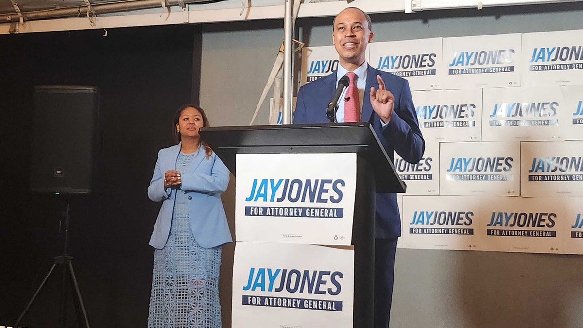 Jay Jones speaks at a podium while wife Mavis Jones stands behind him