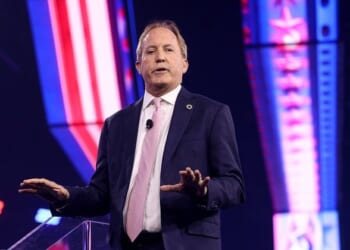 Speaker addressing an audience at a conference, wearing a suit and tie, with colorful stage lighting in the background.