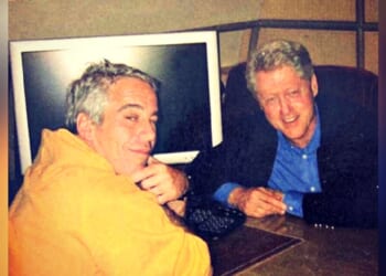 Two men posing together at a desk with a computer in the background, one wearing an orange hoodie and the other in a black jacket and blue shirt.