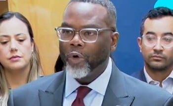 A man in a suit speaks at a press conference, flanked by two individuals, discussing important community issues.