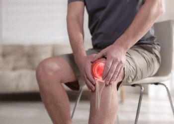 Man sitting on a chair, holding his knee in pain, indicating a potential knee injury or discomfort in a bright indoor setting.