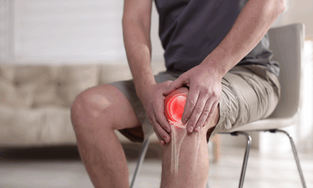 Man sitting on a chair, holding his knee in pain, indicating a potential knee injury or discomfort in a bright indoor setting.