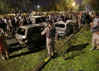 Crowd gathers around damaged vehicles at night, following an incident, with security personnel present and emergency response visible.