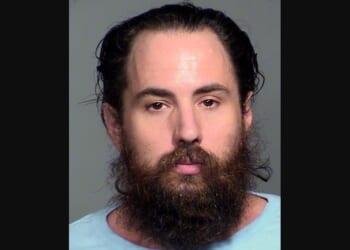 Mugshot of a man with long hair and a full beard, wearing a light blue shirt against a neutral background.