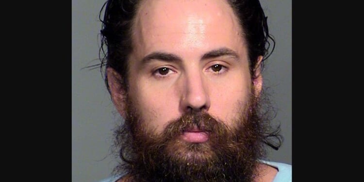 Mugshot of a man with long hair and a full beard, wearing a light blue shirt against a neutral background.