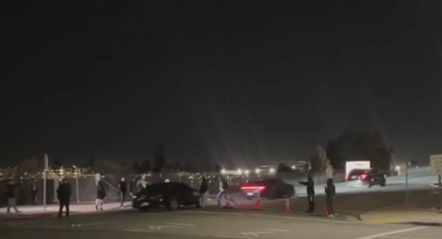 Crowd gathered at night near parked cars on a dimly lit street, with city lights visible in the background.