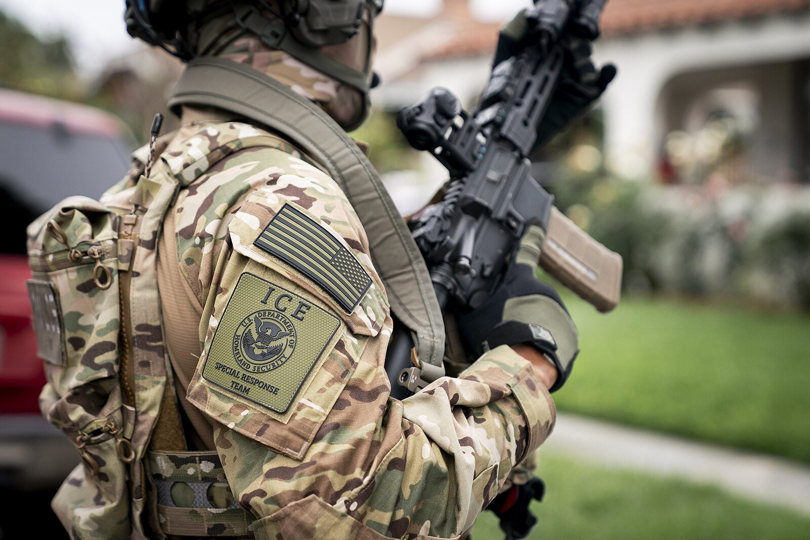 ICE special response team member in tactical gear holding a firearm, showcasing a camouflage uniform and agency patches.