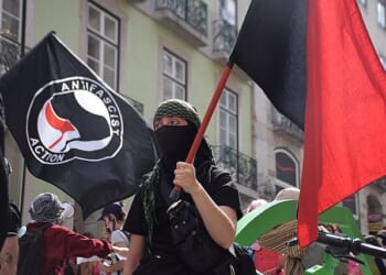 Person in a black mask and green headscarf holding a red flag at an anti-fascist rally with banners in the background.