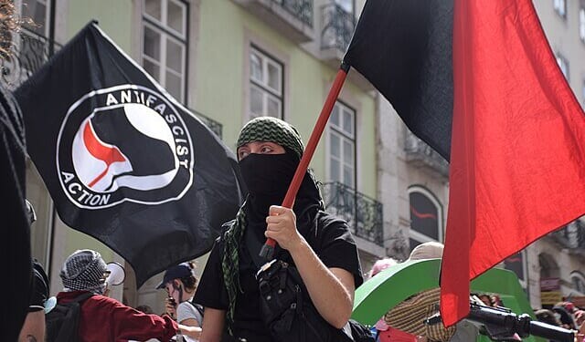 Person in a black mask and green headscarf holding a red flag at an anti-fascist rally with banners in the background.