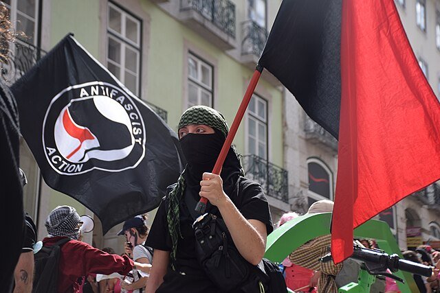 Person in a black mask and green headscarf holding a red flag at an anti-fascist rally with banners in the background.