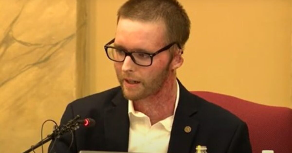 Man with facial scars and glasses speaking at a podium during a formal meeting, emphasizing a serious topic.