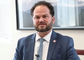 Man in a suit sitting in an office, with a mountain landscape photo in the background, looking directly at the camera.
