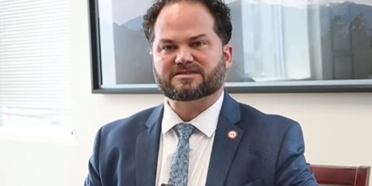 Man in a suit sitting in an office, with a mountain landscape photo in the background, looking directly at the camera.