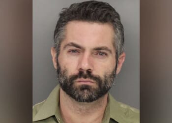 Mugshot of a man with a beard and short hair, wearing a green shirt, looking directly at the camera against a gray background.