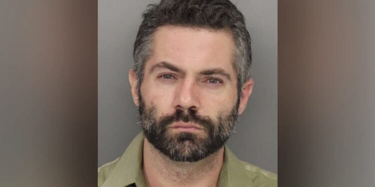 Mugshot of a man with a beard and short hair, wearing a green shirt, looking directly at the camera against a gray background.