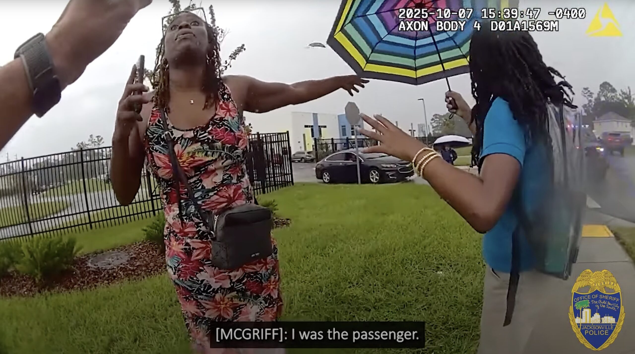 Two women interact during a police encounter, one holding an umbrella and expressing her perspective while the other gestures, set in a grassy area with a fence in the background.