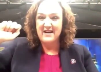 Woman in a blazer passionately speaking with a raised fist, expressing strong emotions in a broadcast studio setting.