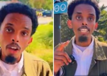 A man with curly hair and a beard speaks passionately outdoors, gesturing with his hand against a backdrop of greenery and urban scenery.