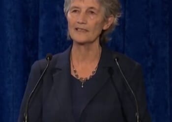 Woman speaking at a podium with a wooden sign in Irish, set against a blue backdrop, conveying a message to an audience.