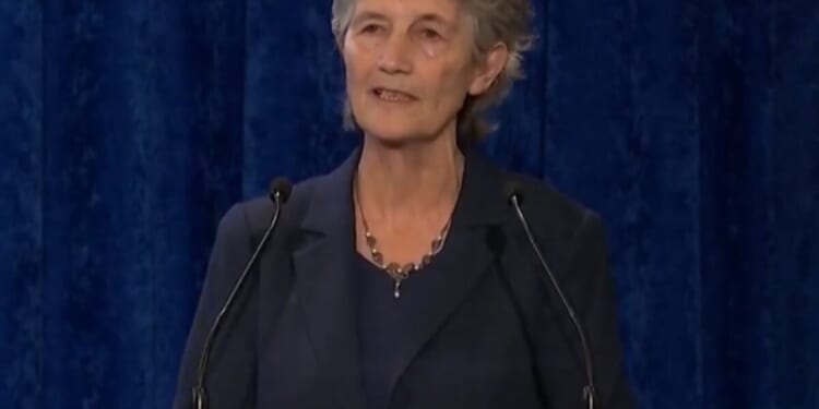 Woman speaking at a podium with a wooden sign in Irish, set against a blue backdrop, conveying a message to an audience.
