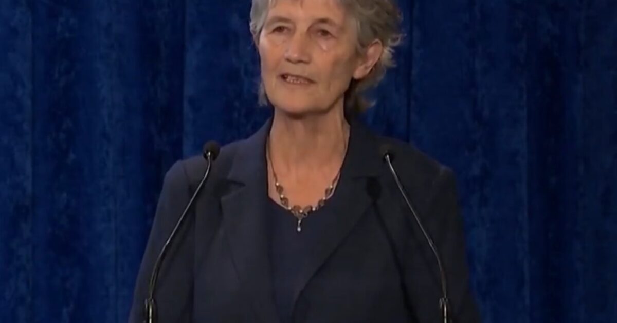 Woman speaking at a podium with a wooden sign in Irish, set against a blue backdrop, conveying a message to an audience.