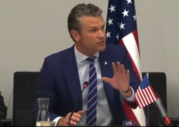 Man in a suit speaking during a formal meeting, with a U.S. flag in the background and a glass of water on the table.