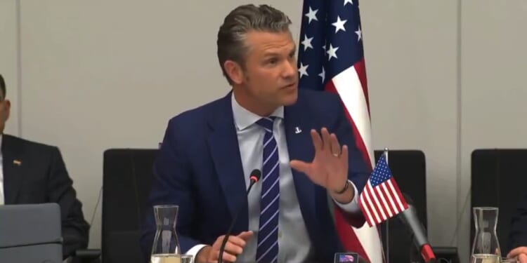 Man in a suit speaking during a formal meeting, with a U.S. flag in the background and a glass of water on the table.