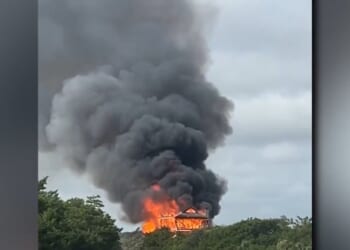 A house engulfed in flames emits thick black smoke against a cloudy sky, surrounded by greenery.