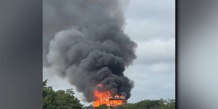 A house engulfed in flames emits thick black smoke against a cloudy sky, surrounded by greenery.