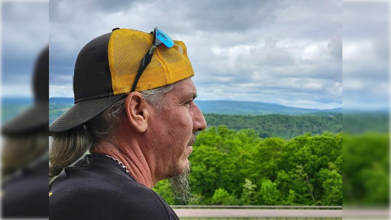 Man wearing a yellow and black cap gazes thoughtfully at a scenic landscape of green hills under a cloudy sky.