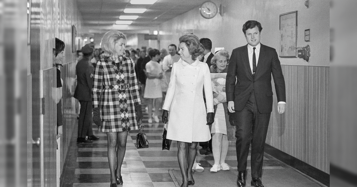 Black and white photo of three well-dressed individuals walking through a hospital corridor, with nurses and visitors in the background.