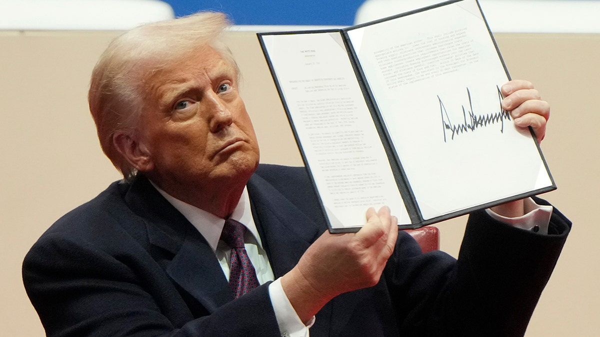 Donald Trump wearing a dark suit and looking serious as he holds up an executive order he signed