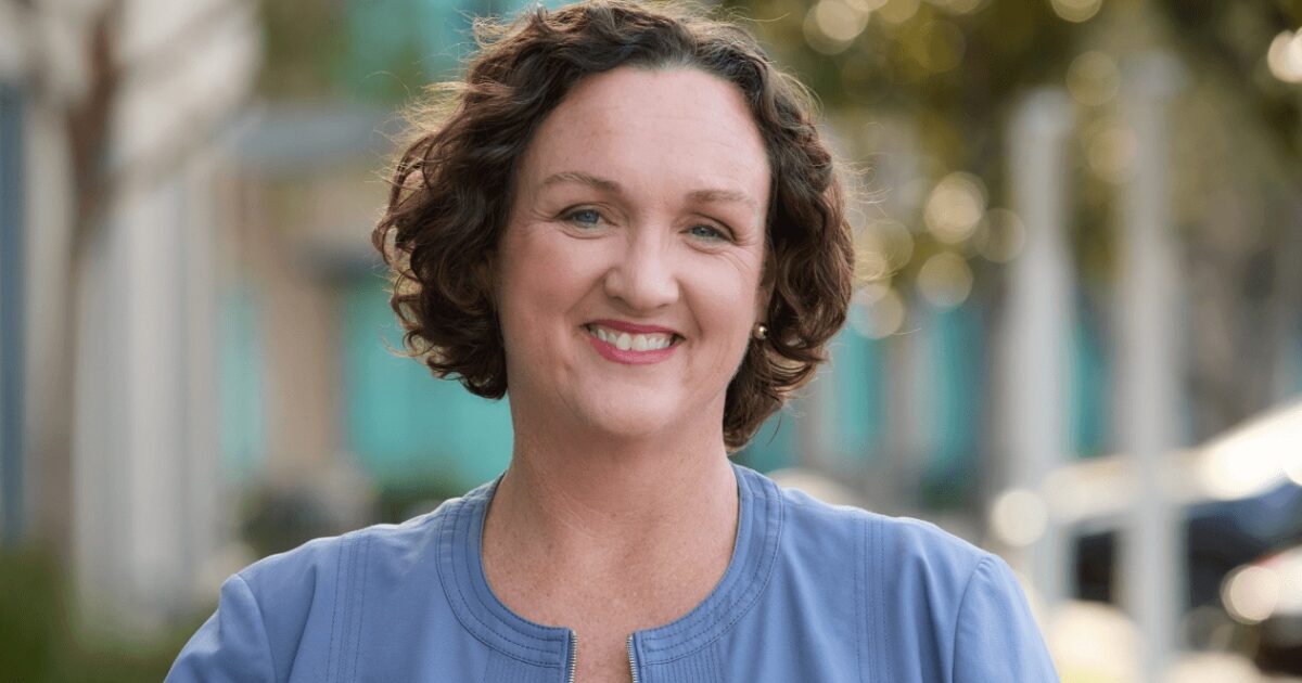 Smiling woman with curly hair wearing a blue jacket, standing outdoors with a blurred background of trees and buildings.