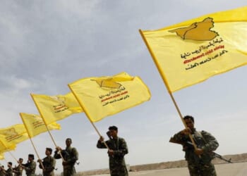 Members of a military group hold yellow flags with symbols and text, representing their organization, against a clear sky backdrop.
