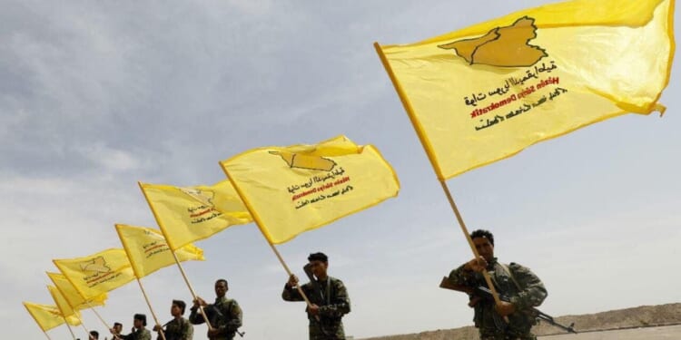 Members of a military group hold yellow flags with symbols and text, representing their organization, against a clear sky backdrop.