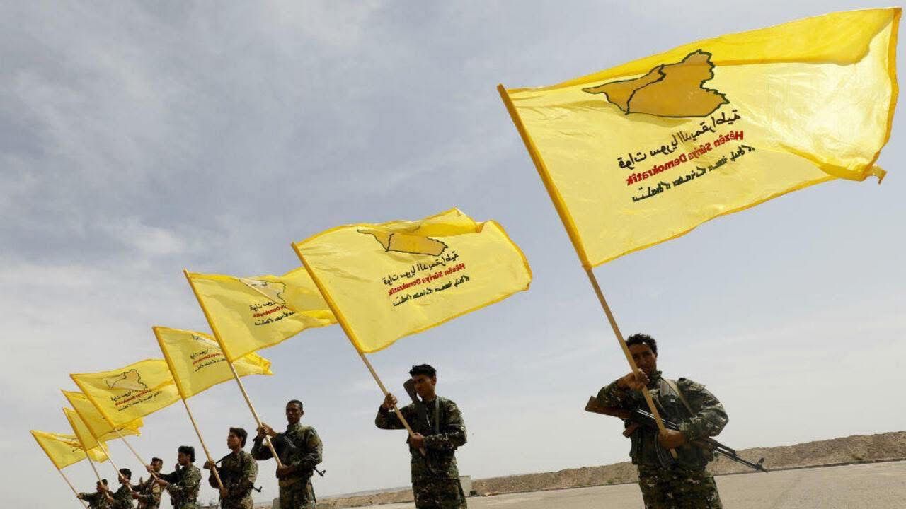Members of a military group hold yellow flags with symbols and text, representing their organization, against a clear sky backdrop.