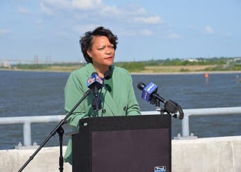 A woman in a green blazer speaks at a podium by a river, with microphones and a scenic background under a clear blue sky.
