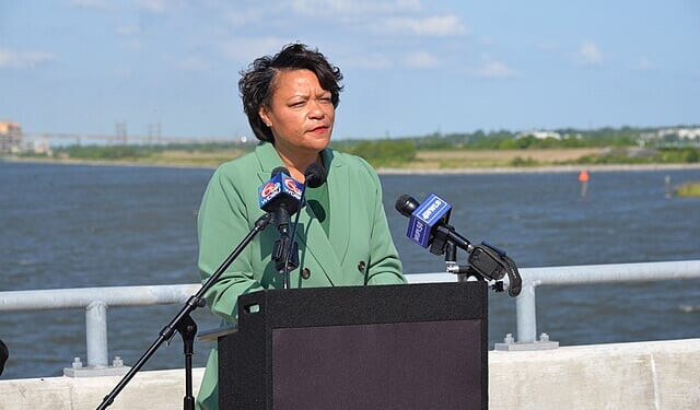 A woman in a green blazer speaks at a podium by a river, with microphones and a scenic background under a clear blue sky.