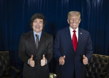 Two men giving thumbs up pose together, one in a suit and tie and the other in a black suit, against a blue curtain backdrop.