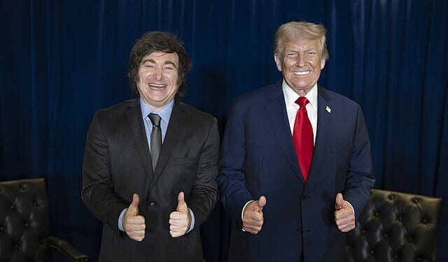 Two men giving thumbs up pose together, one in a suit and tie and the other in a black suit, against a blue curtain backdrop.