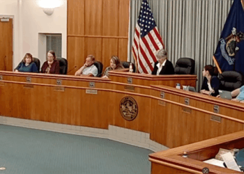 City council meeting in progress with members seated at a wooden table, American flag displayed, discussing local governance issues.